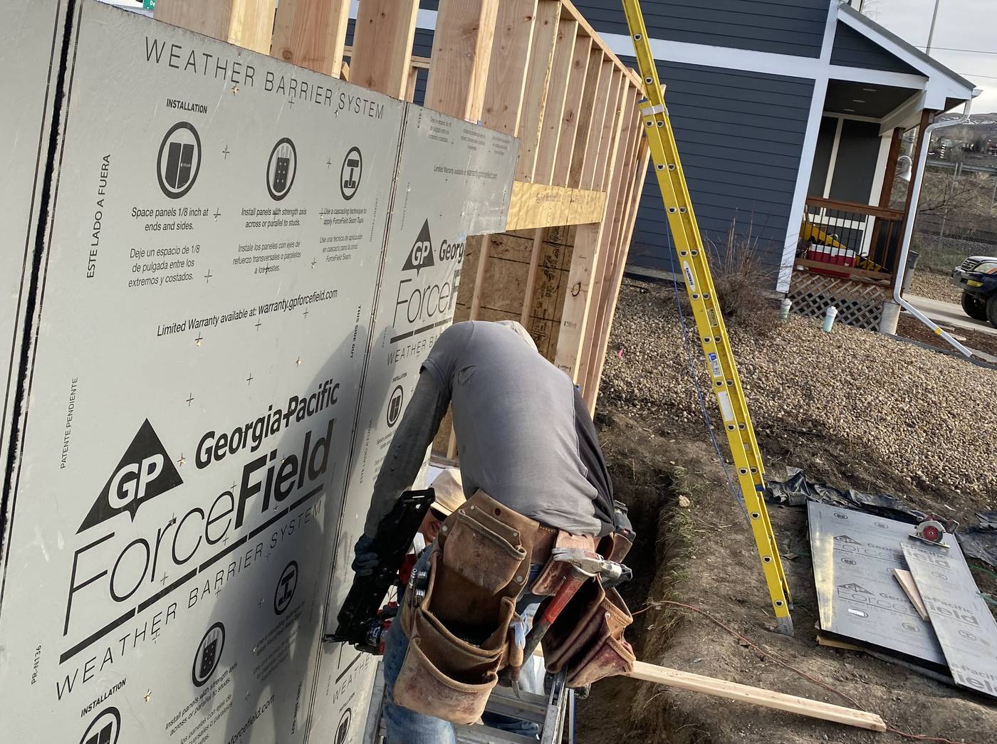 A crew member on a ladder installing weather barrier panels on new wall framing, with a two-story house behind