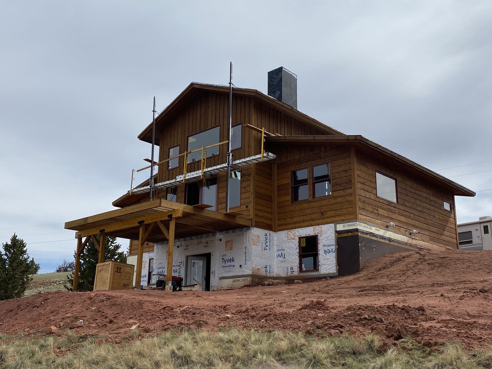 A two-story cedar-sided custom home under construction on a red-dirt mountain site, with a timber-framed porch