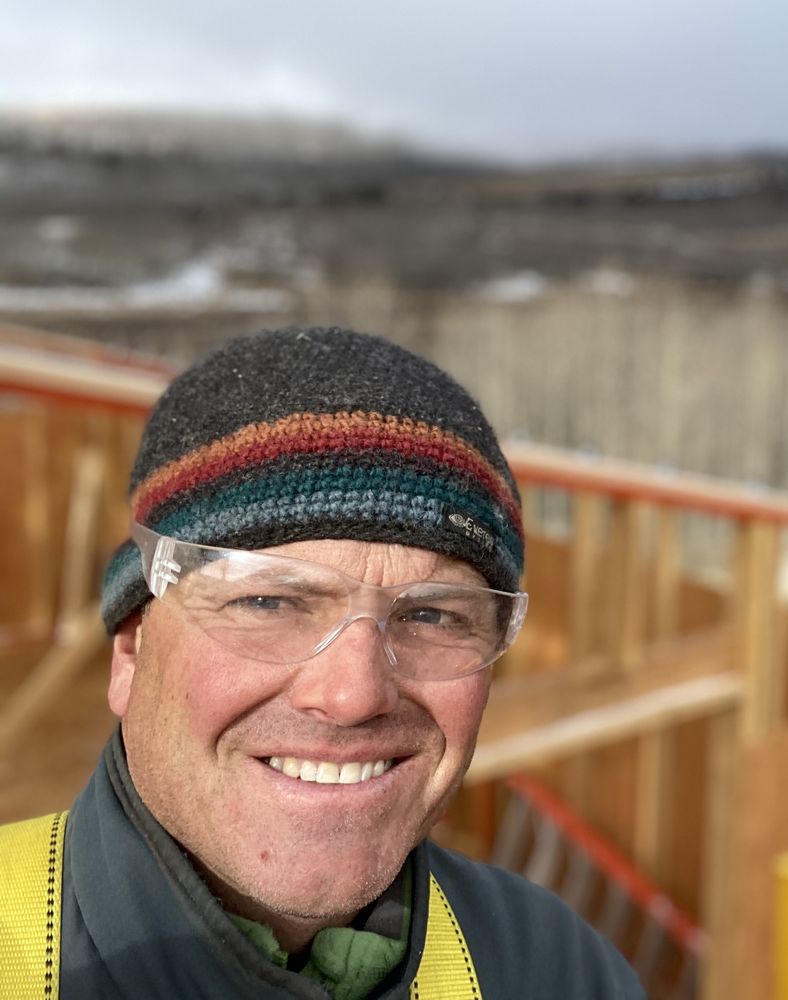 Kurt Ulrich, owner of The Colorado Roofing Company, in safety glasses and a knit hat at a framing site with snowy hills behind